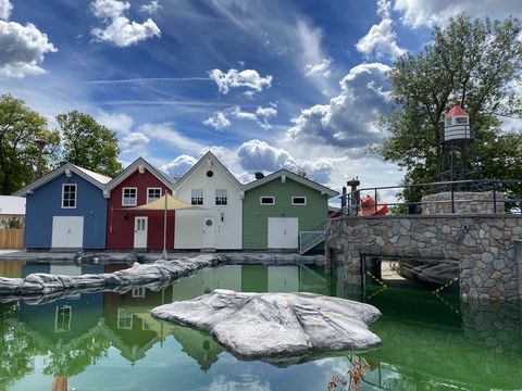 Drei bunte Holzhäuser und ein Leuchtturm spiegeln sich in einem ruhigen Teich unter blauem Himmel.
