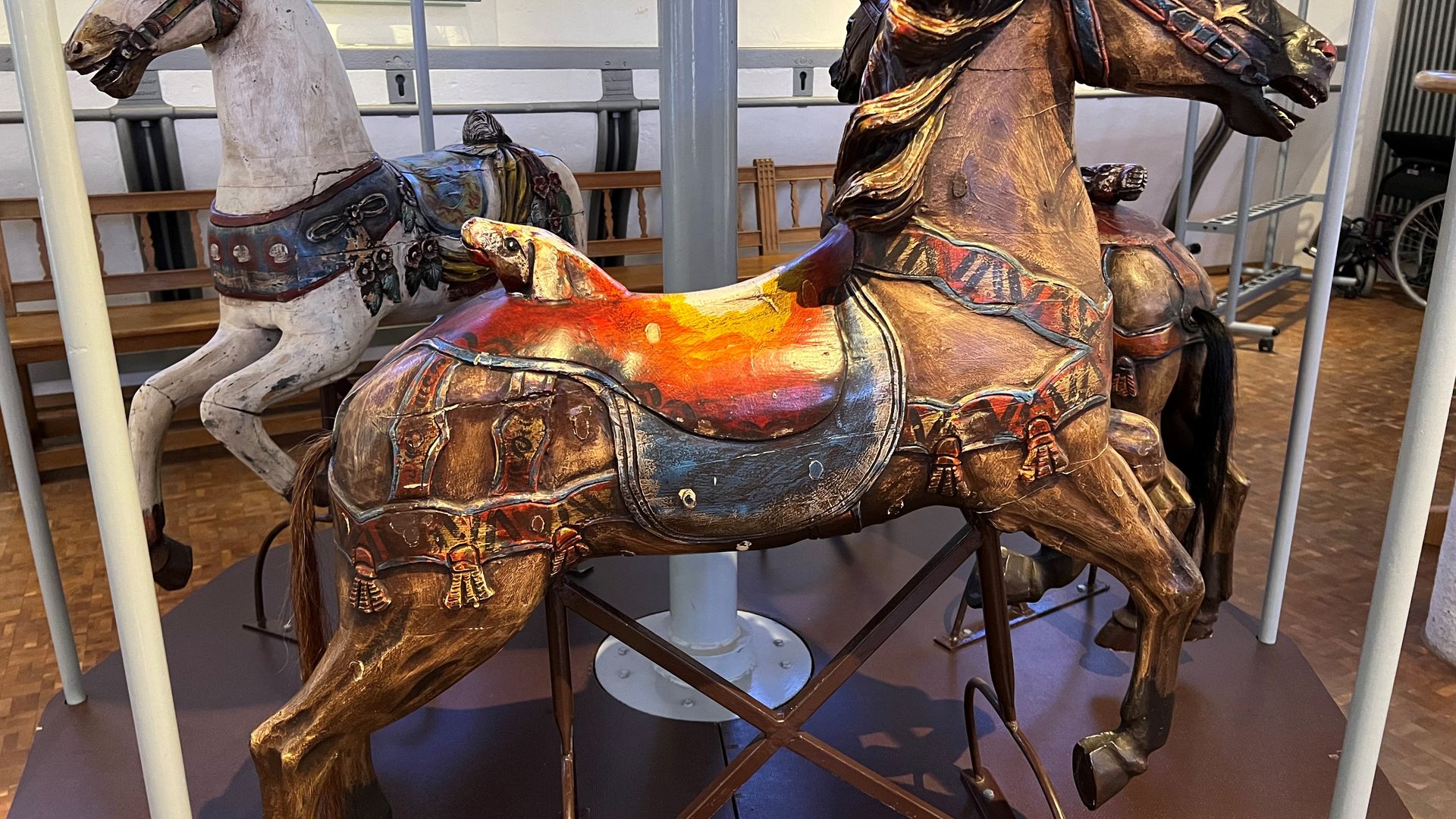 Drei kunstvoll geschnitzte, verzierte hölzerne Karussellpferde stehen in einem musealen Ambiente.